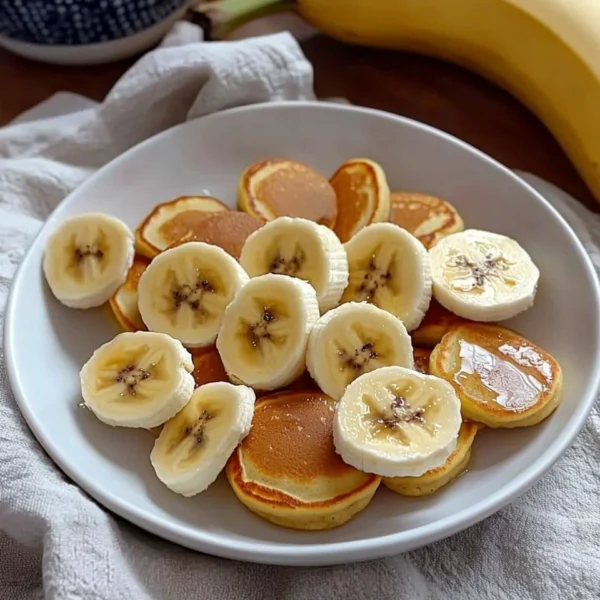 Fluffy banana mini pancakes stacked on a plate, drizzled with syrup.